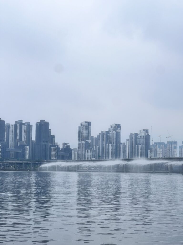 A serene cityscape featuring tall modern skyscrapers reflecting in a calm body of water under a cloudy sky.